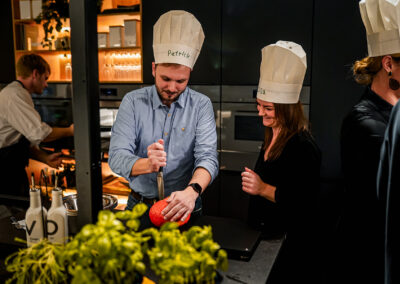 mehrere Leute kochen gemeinschaftlich bei einem Teamevent in der Pioneers Kitchen. Sie haben gute Laune, tragen Kochmützen und schneiden Gemüse.